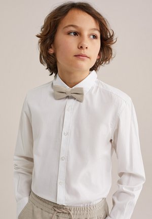 Persona joven con cabello castaño de longitud media, vestida con una camisa blanca abotonada y un lazo beige, con las manos en los bolsillos de los pantalones beige, mirando hacia la derecha.