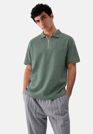 Giovane uomo con capelli scuri e ricci che indossa una polo verde a maniche corte con zip frontale e pantaloni grigi a righe, mani in tasca.