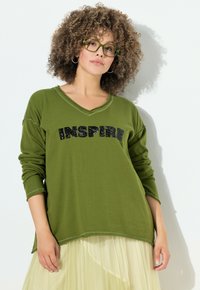 Sudadera verde oliva con cuello en V y mangas largas, que presenta el texto "INSPIRE" en lentejuelas negras. Tejido suave, corte relajado y usada sobre una falda amarilla clara.