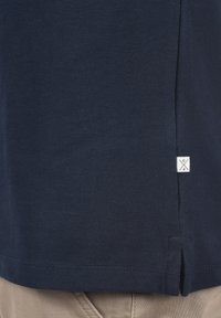 Marineblå polo skjorte i teksturert stoff med splitt på sidene; har en liten hvit etikett med en logo på kanten.