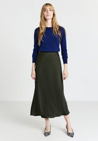 Navyblaues Strickpullover, olivgrüne Satin-Midikleider und schwarz-weiße Zebra-Muster-High Heels. Das Model steht vor einem neutralen Hintergrund.