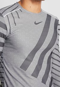 Szara, długosleeve'owa koszulka sportowa Nike z teksturowanymi wzorami i czarnymi paskami. Wykonana z lekkiego, oddychającego materiału.