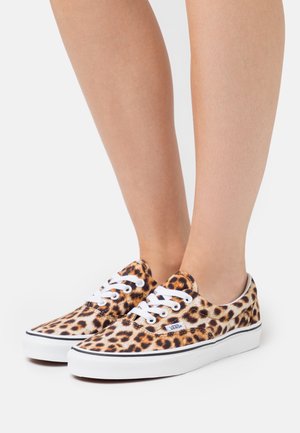 Füße, die niedrige Vans-Sneaker aus Leinwand mit Leopardenmuster und weißen Schnürsenkeln tragen, vor einem weißen Hintergrund.