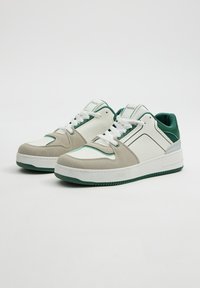 Zapatillas blancas y verdes con parte superior de cuero liso, detalles en ante, cuello acolchado y suela de goma texturizada. Presenta costuras en contraste.