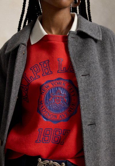 Άτομο που φοράει κόκκινο φούτερ Ralph Lauren με μπλε έμβλημα της Νέας Υόρκης και το κείμενο 1967, φορεμένο κάτω από γκρι παλτό και λευκό πουκάμισο με γιακά.