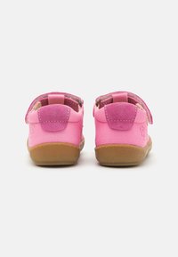 Vaaleanpunaiset lasten kengät, joissa on mokkanahasta ja nahasta valmistettu päällinen, tarranauhat ja kumipohjat. Varustettu pyöreällä nilkalla ja teksturoiduilla yksityiskohdilla.