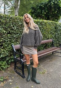 Pull en laine gris confortable avec motif en chevron, jupe beige clair et bottes en caoutchouc vertes. L'arrière-plan présente une haie verte luxuriante et un banc.