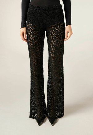 Zwart kanten broek met een bloemenpatroon, uitlopende vorm en hoge taille. Ontworpen met een doorschijnende textuur, gecombineerd met puntige zwarte hakken.