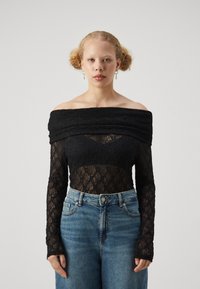 Body in pizzo nero con design off-shoulder, maniche lunghe e dettagli testurizzati su un pannello trasparente, abbinato a jeans blu a vita alta.