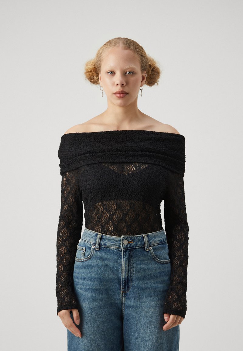 Body in pizzo nero con design off-shoulder, maniche lunghe e dettagli testurizzati su un pannello trasparente, abbinato a jeans blu a vita alta.