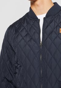 Námořnická prošívaná bomber bunda s zipovým zavíráním, žebrovaným límcem a malou značkovou nášivkou na hrudi. Hladký syntetický materiál.