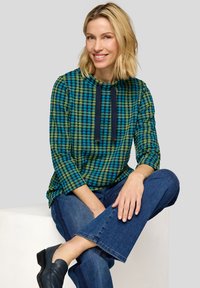 Langärmliges Kapuzenshirt in blau-grün kariertem Muster mit Kordelzug am Hals, kombiniert mit ausgestellten Jeans und schwarzen Schuhen.