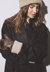 Čierny vlnený kabát s dvojradovým zapínaním, doplnený hnedými koženými rukavicami a béžovou čiapkou s fleecovou podšívkou. Mäkka textúra a kontrastné farby.
