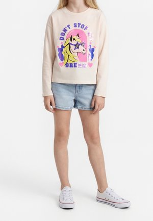 Dziecko w beżowej koszulce z długim rękawem z kolorowym jednorożcem i napisem „Don’t Stop My Dream”, jasnych dżinsowych szortach i białych tenisówkach.