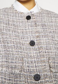 Tvídové sako v béžové a tlumeně fialové barvě s texturovaným povrchem, třepícími se okraji a černými knoflíky. Obsahuje detail malého kapesního prvku.