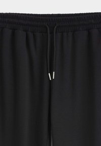 Pantalon de survêtement noir avec une ceinture élastique, doté d'un cordon de serrage avec embouts en métal. Le tissu semble doux avec une texture lisse.