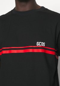 Camiseta de algodón negra con rayas horizontales rojas y logo blanco "GCDS" en el lado izquierdo del pecho. Textura suave con un diseño de cuello redondo.