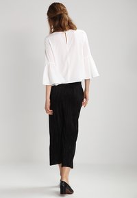 Blusa bianca con maniche a sbuffo e schiena a chiave, abbinata a pantaloni a gamba larga neri a righe verticali e scarpe con tacco nere.