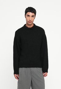 Pull noir en tricot avec un col rond côtelé et des manches longues. Le tissu a une finition texturée. Porté avec un pantalon gris.
