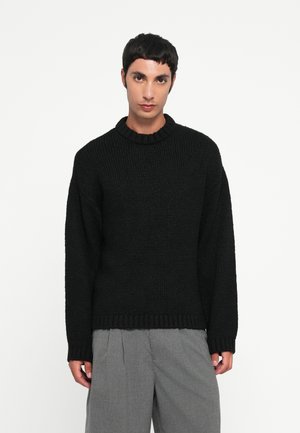 Suéter negro de punto con un cuello redondo acanalado y mangas largas. El material tiene un acabado texturizado. Llevado con pantalones grises.