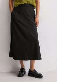 Schwarzer A-Linien-Rock mit glatter Textur, der eine hohe Taille hat und eine Nahtdetail, die vorne entlang verläuft. Kombiniert mit schwarzen Slippers.