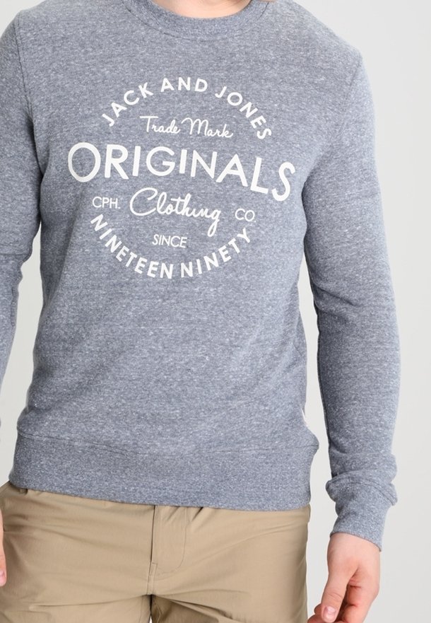 Persona che indossa una felpa grigia Jack and Jones Originals con testo bianco e pantaloni beige su uno sfondo chiaro e uniforme.