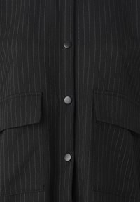 Camicia a righe nere con righe verticali bianche, chiusura con bottoni, due tasche a toppa e una texture di tessuto liscia.