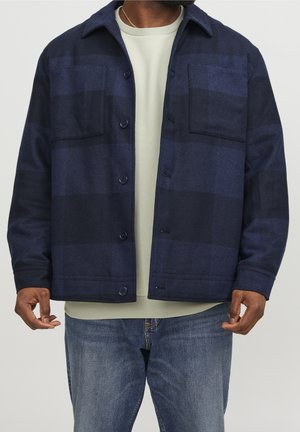 Homme portant une veste à carreaux bleu marine et noir à boutons, une chemise beige clair et un jean bleu, debout devant un fond uni.