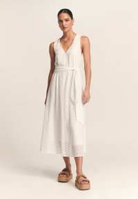 Balta, be rankovių midi suknelė su akių raštu, V formos iškirpte ir audinio diržu. Modelis dėvi rudus platforminius sandalų batus su plačiomis juostomis.