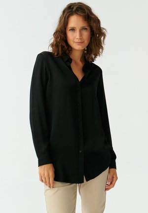 Camicia nera a maniche lunghe con bottoni, realizzata in tessuto morbido, con colletto e vestibilità rilassata, abbinata a pantaloni di colore chiaro.
