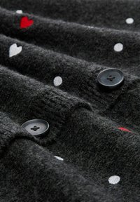 Dunkelgrauer Strickstoff mit schwarzen Knöpfen, der weiße gepunktete Muster und kleine rote und weiße Herzmuster aufweist.
