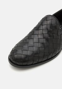 Black woven leather slip-on shoe with a smooth texture, rounded toe, and subtle stitching detail along the edges.