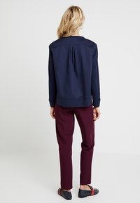 Marineblauer Pullover mit Rundhalsausschnitt, überschnittenen Schultern und gefälteltem Rücken. Kombiniert mit burgunderfarbener Hose und schwarzen Schuhen mit roten Akzenten.