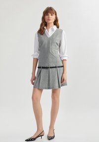 Vestido de cuadros gris con escote en V, falda plisada y un cinturón negro. Llevado sobre una camisa blanca con cuello. Zapatos de tacón negros con adornos.