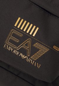 Material textil negru cu un finisaj texturat, având un logo auriu care include "EA7 EMPORIO ARMANI" și accente în dungi.