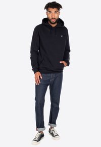 Felpa nera con cappuccio e tasca frontale, abbinata a jeans blu scuri arrotolati sui polsi, e sneakers nere con suola bianca.