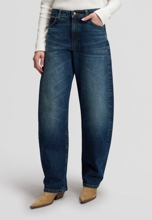 Femme portant un jean bleu foncé ample taille haute, un top blanc à manches longues et des bottines à talons pointus couleur camel, debout avec une main dans la poche.