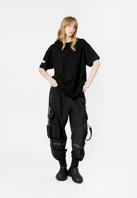 Camiseta negra de gran tamaño con un sutil detalle de texto, combinada con pantalones cargo negros que cuentan con cremalleras y bolsillos. Llevada con zapatos negros texturizados.