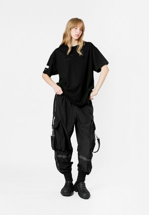 Camiseta negra de gran tamaño con un sutil detalle de texto, combinada con pantalones cargo negros que cuentan con cremalleras y bolsillos. Llevada con zapatos negros texturizados.