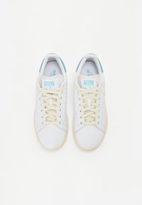 Hvide lædersneakers med lyse beige snørebånd og såler, der har en turkise detalje ved hælen og blå branding på tungen.