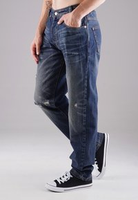 Dunkelblaue Jeans mit lockerer Passform, die verschlissene Details und Risse aufweisen. Kombiniert mit schwarzen Low-Top-Sneakern mit weißen Akzenten.