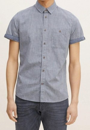 Mand iført kortærmet lyseblå skjorte med brystlomme og mørkeblå jeans, stående med armene afslappet ned langs siden.