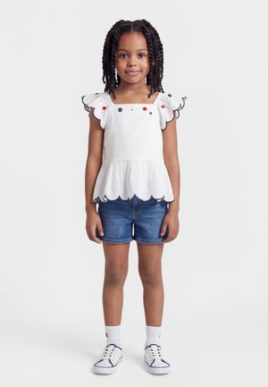 Dievča s vrkočmi, ktoré má na sebe bielu blúzku so zaoblenými okrajmi a kvetinovými detailmi, rifľové šortky, biele ponožky a biele tenisky stojace na jednoduchom pozadí.
