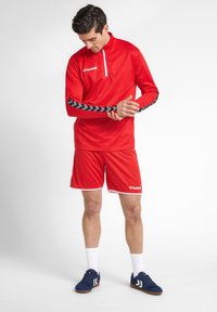 Rood sportoutfit bestaande uit een longsleeve top met halve rits en zwarte chevronstrepen, gecombineerd met bijpassende shorts, beide met witte accenten.