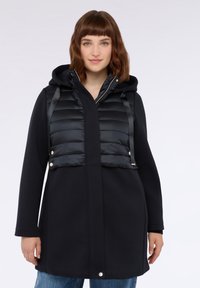 Schwarzer Mantel mit einer gesteppte Vorderseite, der eine Kapuze und einen geraden Schnitt hat. Der gesteppte Bereich hat eine gesteppten Textur und goldfarbene Hardware-Akzente.