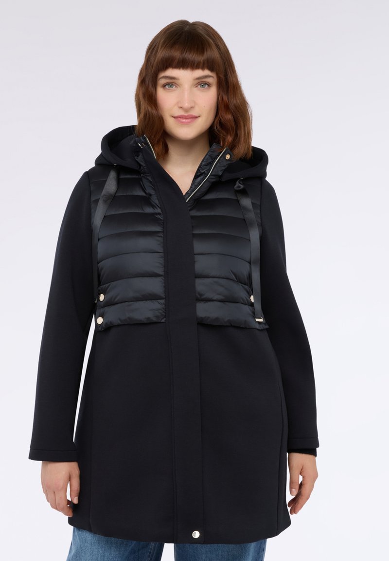 Schwarzer Mantel mit einer gesteppte Vorderseite, der eine Kapuze und einen geraden Schnitt hat. Der gesteppte Bereich hat eine gesteppten Textur und goldfarbene Hardware-Akzente.