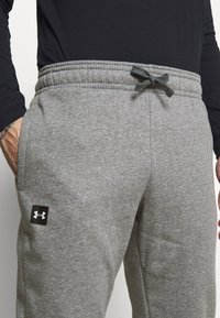 Szare dresy z elastyczną talią, wewnętrznym sznurkiem ściągającym oraz bocznymi kieszeniami. Na lewej udzie znajduje się czarne logo Under Armour.