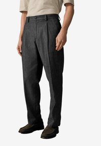 Pantalones gris oscuro con pliegues, fabricados en tela suave, que cuentan con un cierre de botón y bolsillos laterales, combinados con zapatos oscuros.