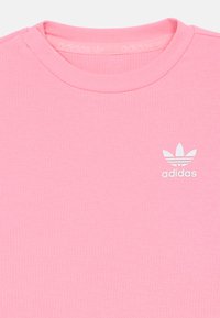 T-shirt rosa com textura, gola redonda, apresentando o logótipo trefoil branco da Adidas e texto no lado esquerdo do peito.