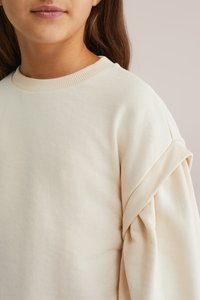 Felpa beige chiaro con colletto a girocollo a costine e maniche piegate. Tessuto in cotone morbido con una finitura leggermente strutturata.